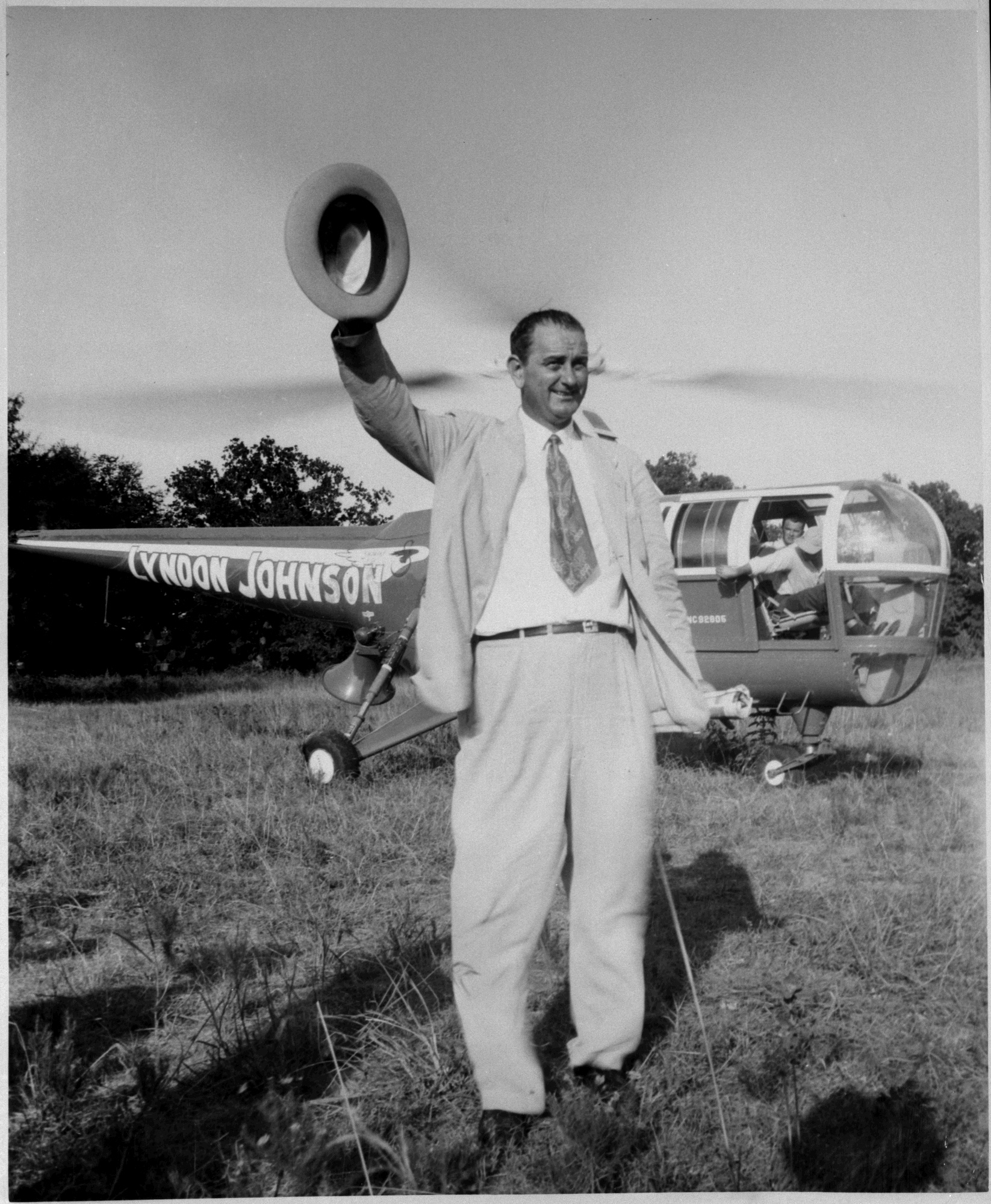 LBJ stands in front of a helicopter with his name on it and waves his hat.